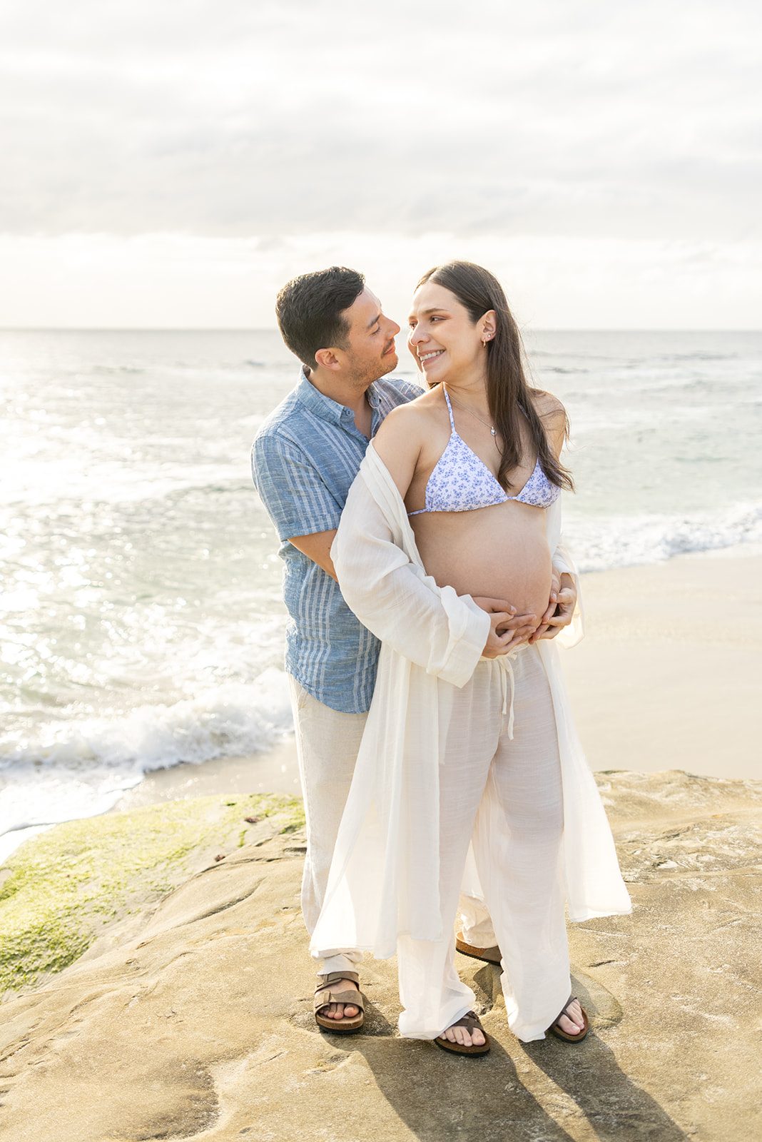 Expecting couple hold the bump together on the sandstone at a San Diego beach maternity session