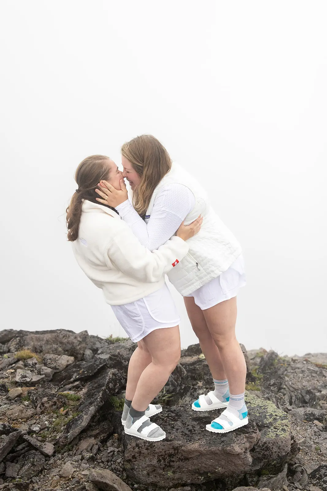 Two brides kiss on dark volcanic rock in heavy fog during their Mt. Rainier elopement ceremony on Burroughs Mountain Trail