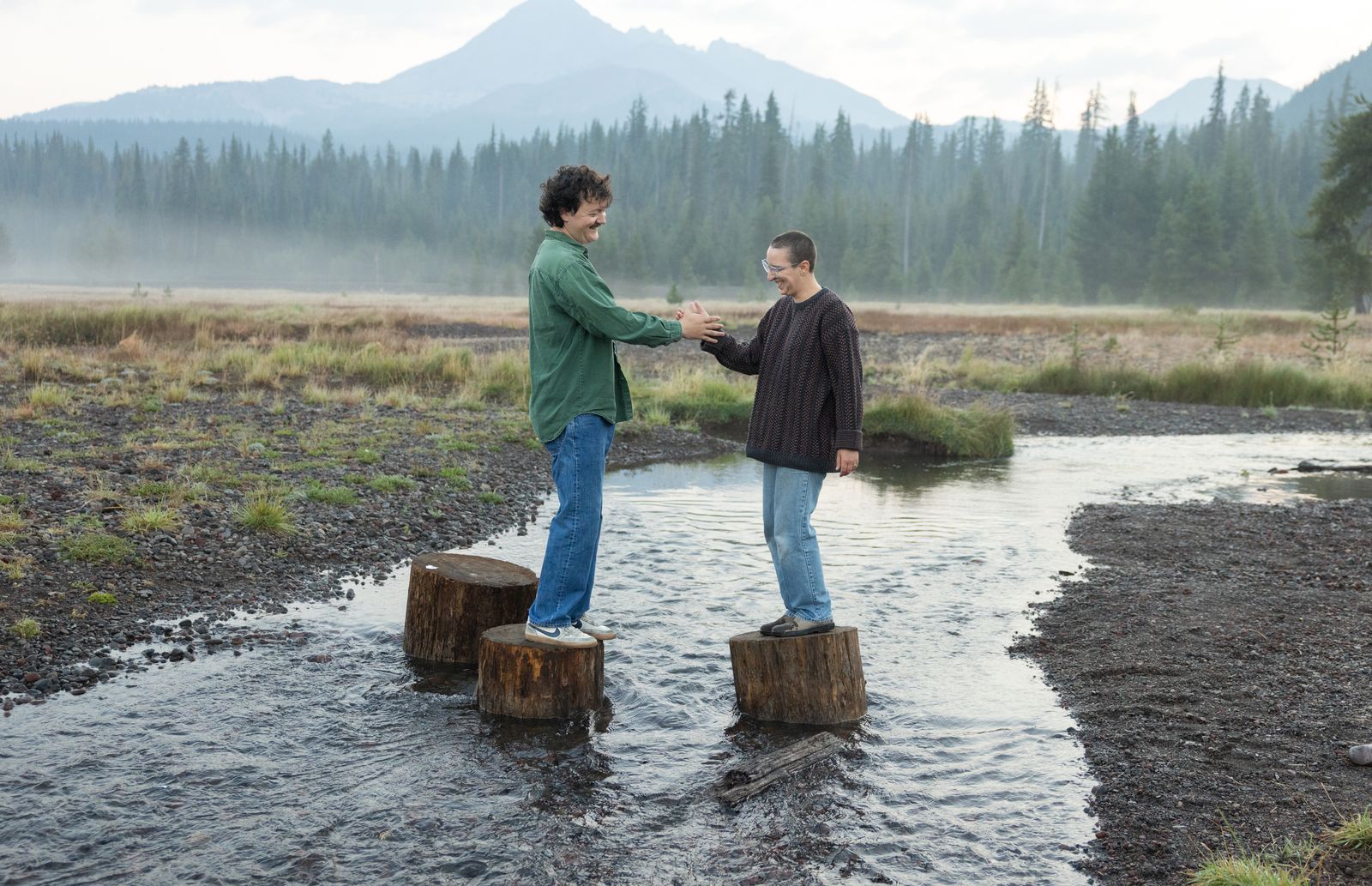 Couple hold hands standing on stumps in a shallow river at sunrise near Sparks Lake