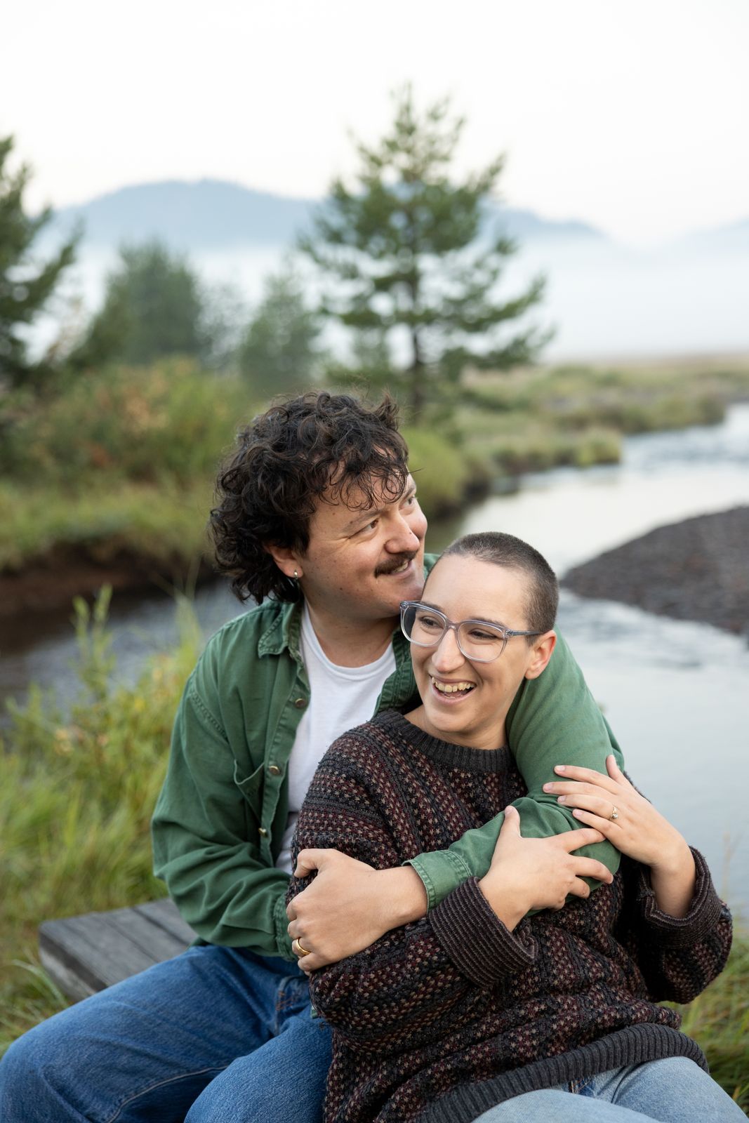 Engaged couple hold each other laughing beside a river in a Central Oregon meadow