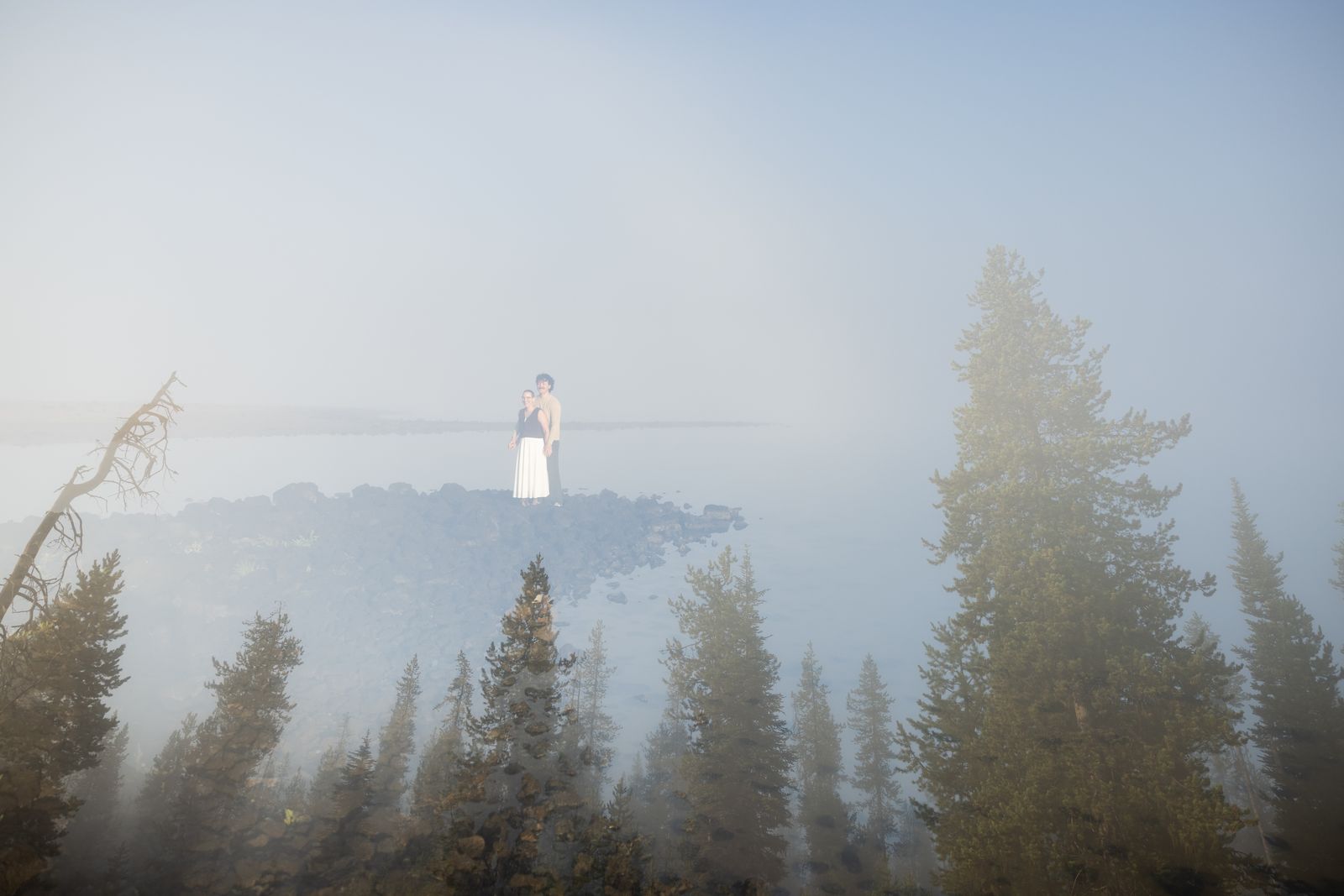 A couple stands far out on the shoreline of Sparks Lake surrounded by thick morning fog